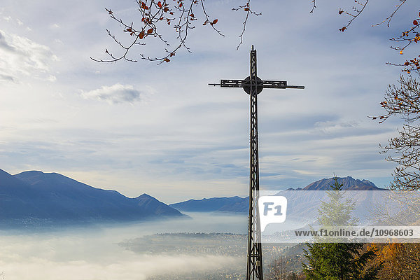 'A cross in the foreground and fog over a city and alpine lake; Locarno  Ticino  Switzerland'