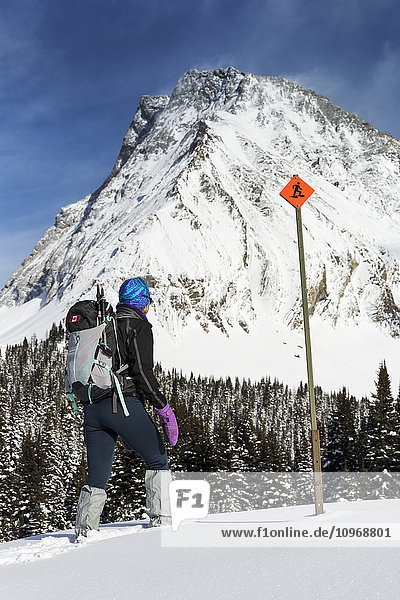 'Female snowshoer on snow covered hillside with snow covered mountains and trees in the background with snowshoe sign and blue sky with clouds; Kananaskis Country  Alberta  Canada'