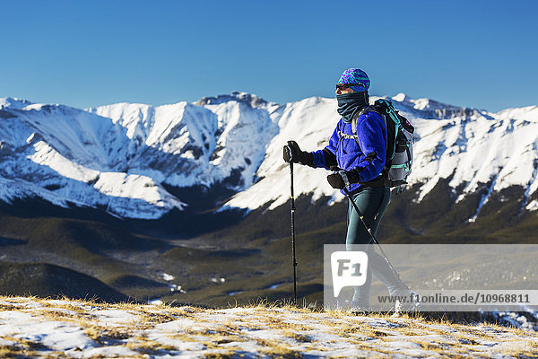 'Female hiker walking on top of a snow covered ridge with foothills below and snow covered mountain range and blue sky in the background; Kananaskis Country  Alberta  Canada'