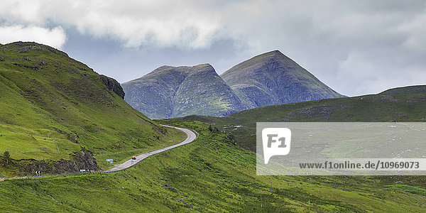 Eine Straße  die sich durch die Berge schlängelt  mit zwei Berggipfeln unter einem wolkenverhangenen Himmel in den Highlands; Schottland