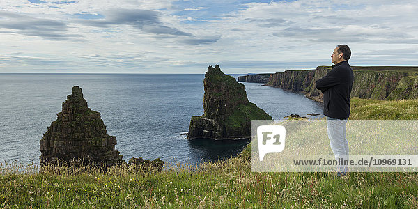 'Man standing and looking out over the North Sea with rugged cliffs and sea stacks along the coastline; Scotland'
