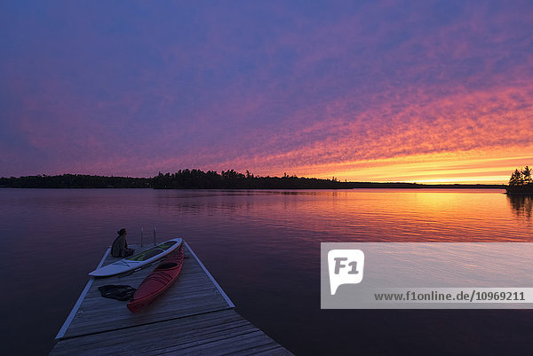 Eine junge Frau sitzt am Ende eines Stegs mit Kajaks und beobachtet den Sonnenuntergang über einem See; Ontario  Kanada'.
