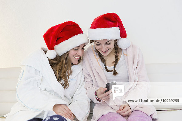 Zwei Schwestern im Teenageralter  die Weihnachtsmannmützen  Schlafanzüge und Gewänder tragen und zusammen auf einer Couch sitzen und auf ein Handy schauen; Bonn  Nordrhein Westfalen  Deutschland'.