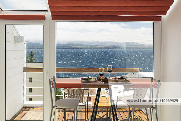 Ein Mittagstisch mit zwei Gläsern Rotwein ist vor dem Balkonfenster einer hellen Wohnung mit Blick auf einen See gedeckt; Bariloche  Argentinien'.