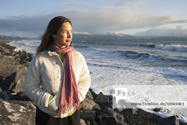 Woman standing along the rocky shoreline of Homer Spit at sunset in winter  Southcentral Alaska