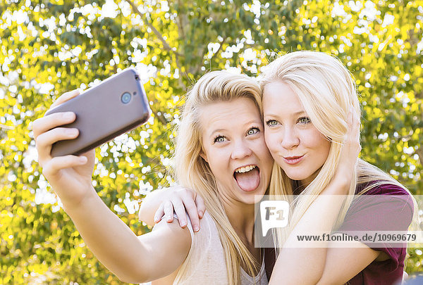 Zwei Schwestern  die sich im Herbst in einem Stadtpark vergnügen  Selfies von sich machen und lustige Grimassen schneiden; Edmonton  Alberta  Kanada'.