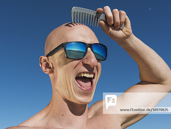 'A guy with a bald head holds a comb over his hairless head while wearing sunglasses and a big smile; Tarifa  Cadiz  Andalusia  Spain'