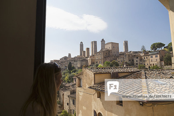 Eine Touristin schaut aus dem Fenster auf das ummauerte mittelalterliche Dorf San Gimignano; San Gimignano  Provinz Siena  Toskana  Italien'.