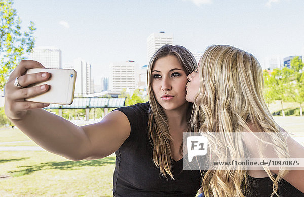 Zwei Freundinnen machen Selbstporträts von sich im Freien in einem Stadtpark  wobei eine Freundin die andere auf die Wange küsst; Edmonton  Alberta  Kanada'.