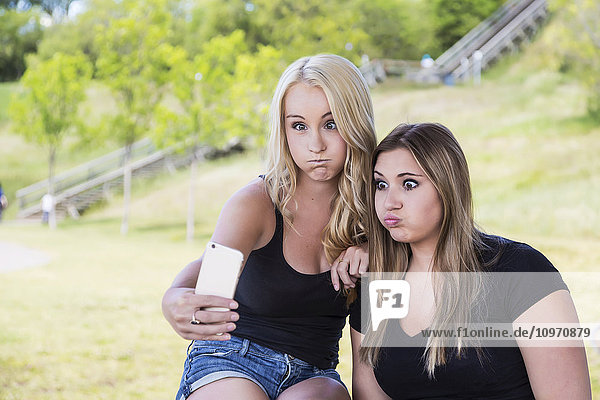 Zwei Freundinnen machen Selfies von sich und schneiden Grimassen in einem Park; Edmonton  Alberta  Kanada'.