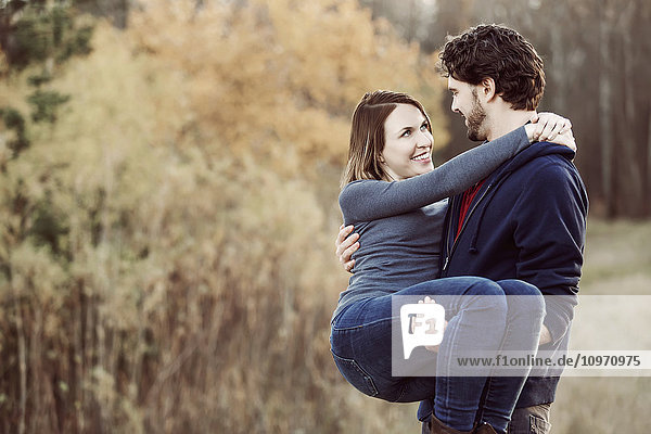 Ein junger Mann nimmt seine Freundin auf den Arm  während er im Herbst in einem Stadtpark spazieren geht; Edmonton  Alberta  Kanada'.
