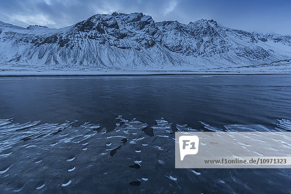Schneebedeckte Landschaft entlang der Ostküste Islands  während ein Strom sanft vorbeifließt und sich Eis entlang des Ufers bildet; Island