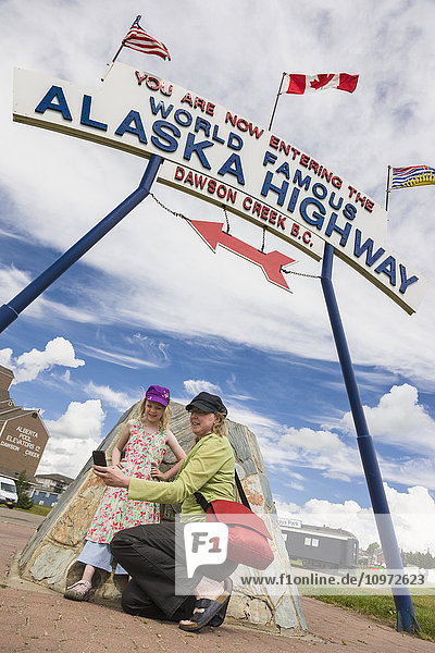 'Mother and daughter take a selfie underneath the ''World Famous Alaska Highway'' sign  Dawson Creek  British Columbia  Canada'