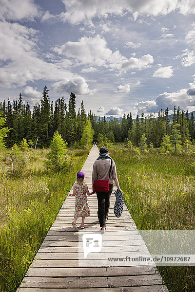 A mother and daughter walk down a boardwalk through a wetland surrounded by evergreen trees  Liard River Hot Springs Provincial Park  British Columbia  Canada  Summer