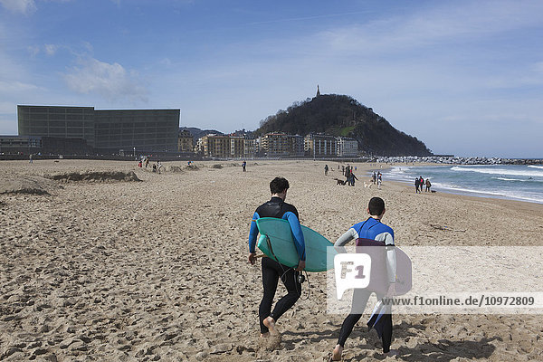 Zwei Surfer in Neoprenanzügen  die mit den beiden Glaswürfeln des Kursaals  eines Kulturzentrums  neben dem Rio Urumea und dem Strand Zurriola im Stadtteil Gros auf die Wellen zugehen; San Sebastian  Spanien'.