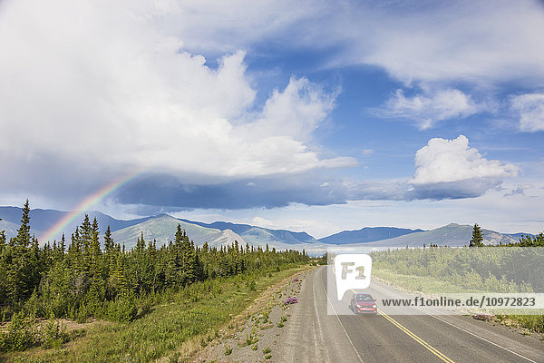 A car drives on the Alaska Highway adjacent to Kluane Lake with atmospheric rain clouds and a Rainbow in the background  Yukon Territory  Canada  Summer