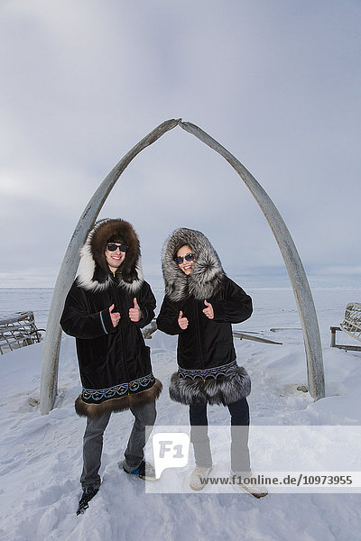 Männliche und weibliche einheimische Jugendliche tragen traditionelle Pelzparkas  während sie neben dem Browerville Walebone Arch stehen  Barrow  North Slope  Arctic Alaska  USA  Winter'.