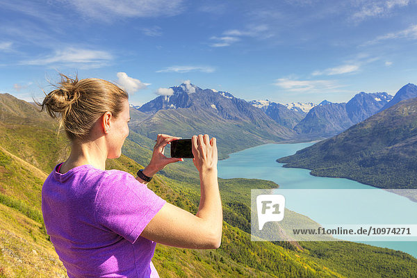 'Female hiker using a smartphone to photograph Eklutna Lake and the Chugach Mountains from the Twin Peaks Trail  Chugach State Park  Southcentral Alaska  Summer'