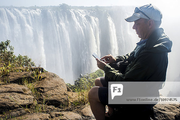 Mann mit Smartphone vor dem Wasserfall der Victoriafälle; Victoria Falls  Simbabwe'.