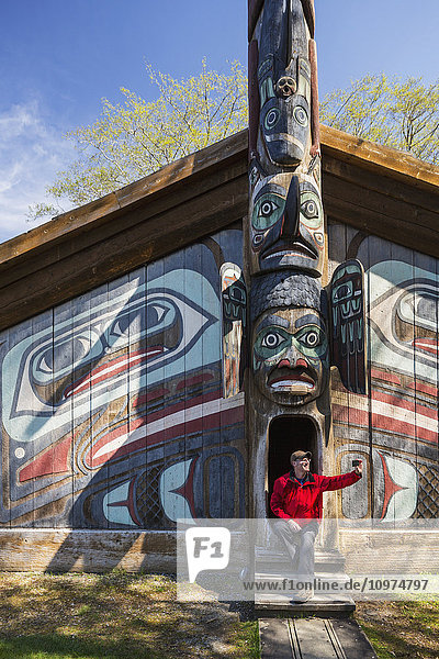 Mann sitzt mit Smartphone auf den Stufen eines Tlingit-Clanhauses  Totem Bight Historic State Park  Ketchikan  Südost-Alaska  USA  Frühling