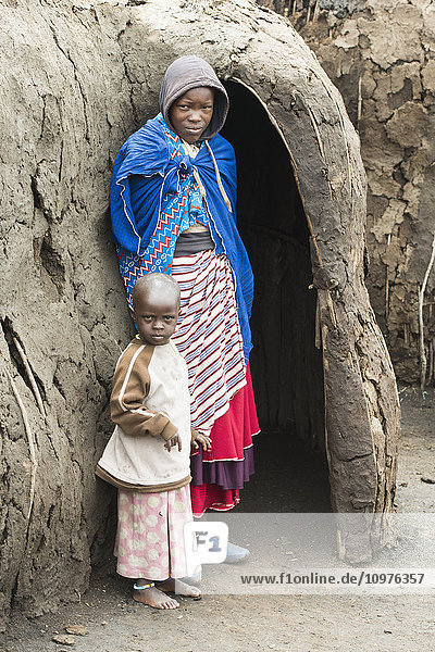 'Maasai mother and child stand at entrance to traditional mud hut  Ngorongoro Conservation Area; Tanzania'