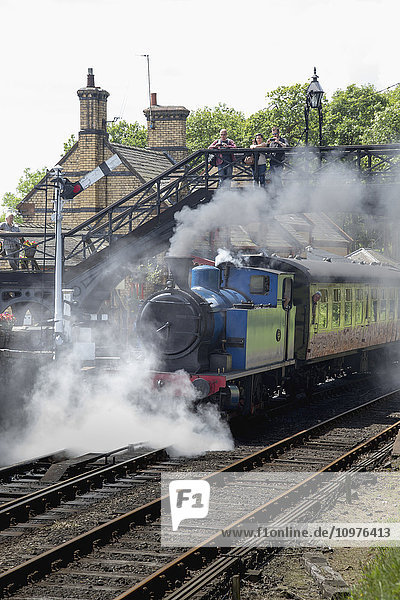 Dampf aus einem Personenzug  der die Gleise hinunterfährt  während Menschen von einem Steg über den Gleisen zusehen; Cumbria  England'.