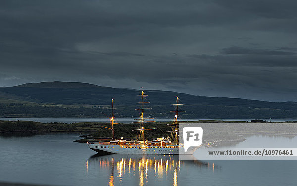 Ein großes weißes Boot mit beleuchteten Masten auf dem ruhigen Wasser entlang der Küste in der Abenddämmerung; Tobermory  Isle of Mull  Argyll und Bute  Schottland'.