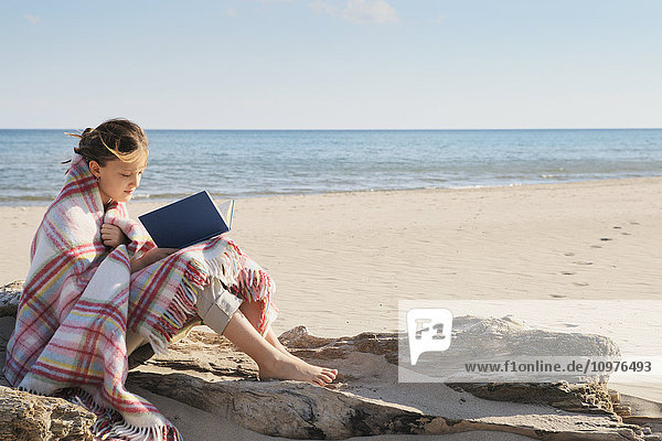 Mädchen  in eine Decke eingewickelt  sitzt auf einem Baumstamm am Strand und liest ein Buch; Toronto  Ontario  Kanada'.