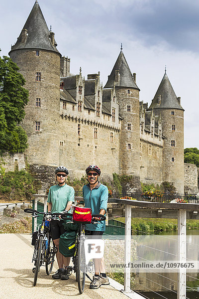 Radfahrendes Paar mit Fahrrädern entlang eines Uferweges und einer alten Steinburg mit großen Türmen im Hintergrund; Josseline  Bretagne  Frankreich'.