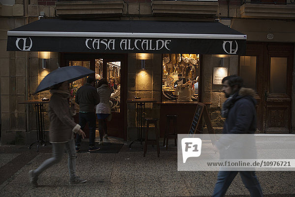 Fußgänger und Menschen  die in der Abenddämmerung eine Bar in der Calle Major betreten  eine Frau trägt einen Regenschirm; San Sebastian  Spanien'.