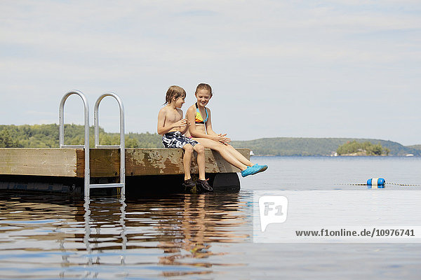 'Girl and boy sitting at end of dock on Mara Lake; Ontario  Canada'