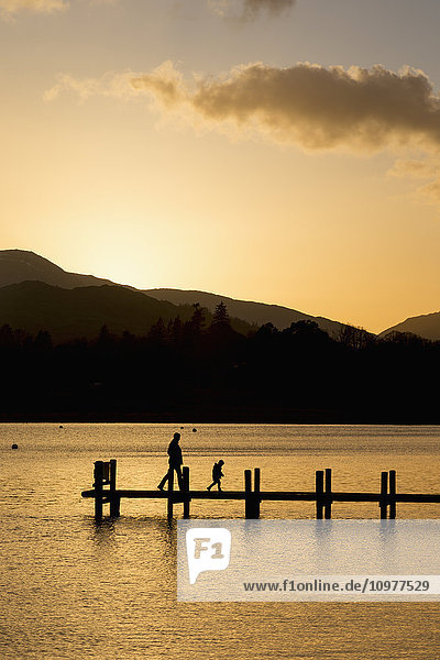 Silhouette eines Elternteils und eines Kindes  die bei Sonnenuntergang auf einem Steg an einem ruhigen See spazieren gehen; South Lakeland District  England'.