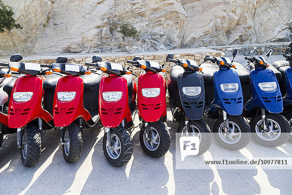 Eine farbenfrohe Reihe von Motorrollern  aufgereiht bei einer Vermietungsagentur neben dem Hafen für Kreuzfahrtschiffe  die Touristen mieten können; Insel Mykonos  Griechische Inseln  Griechenland'.