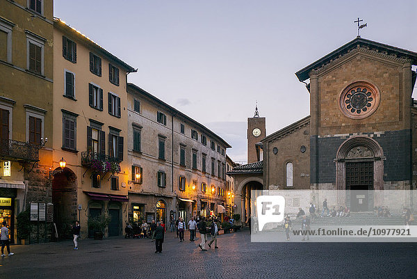 'Pedestrians walk in a town square with shops and a church at dusk; Orvieto  Umbria  Italy'