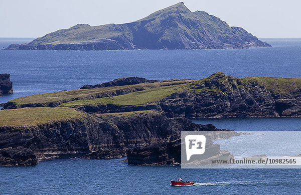 'A small red boat traveling along the coastline with a view of a rugged small island; Portmagee  County Kerry  Ireland'