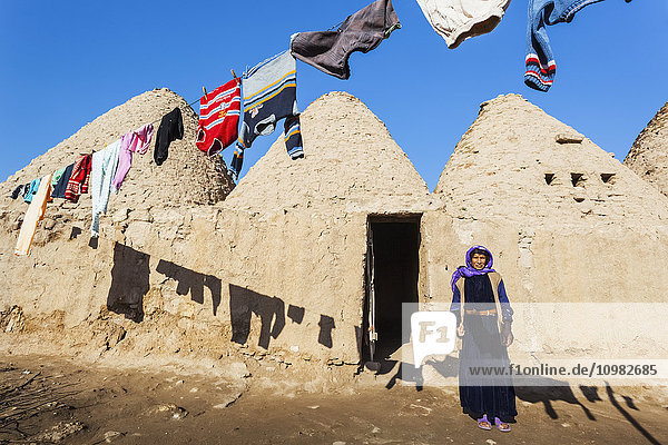 Eine Frau steht vor der Tür ihrer primitiven Behausung im Schatten der auf der Wäscheleine hängenden Kleidung; Harran  Türkei