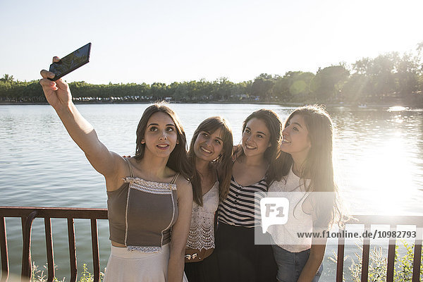 Vier Freunde stehen im Sonnenlicht vor dem Wasser und nehmen Selfie mit dem Smartphone.