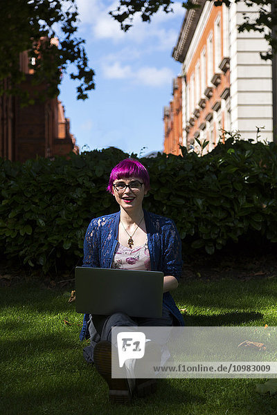 Junge Frau mit gefärbtem Haar auf einer Wiese sitzend mit Laptop