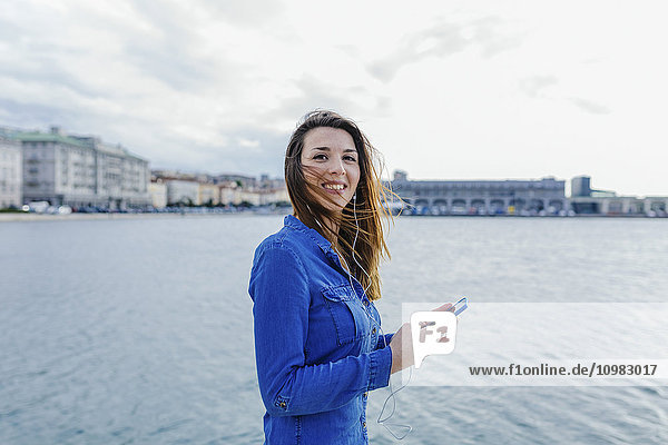 Italy  Trieste  smiling young woman listening to music at the waterfront