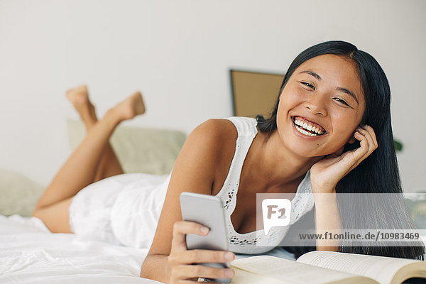 Laughing young woman lying in bed holding cell phone