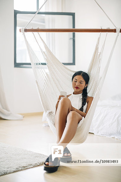 Young woman sitting in hammock using cell phone