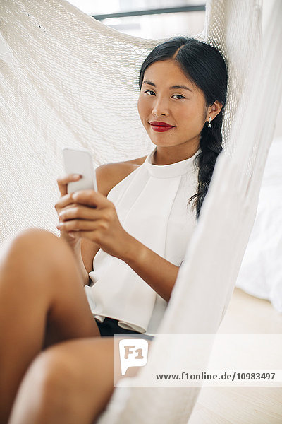 Portrait einer jungen Frau in Hängematte mit Handy