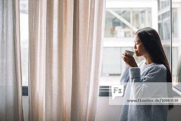 Junge Frau mit Tasse Kaffee vor geöffnetem Fenster