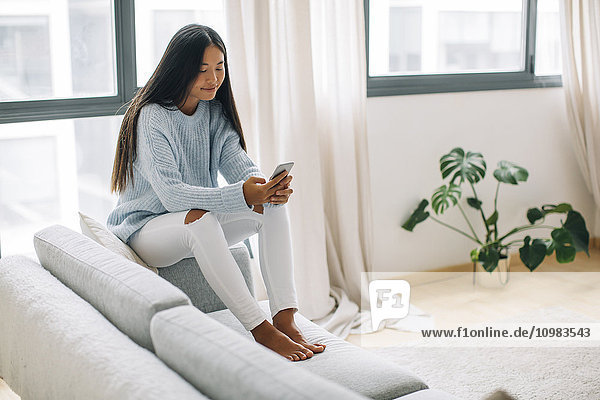 Junge Frau sitzt auf der Couch im Wohnzimmer mit Smartphone