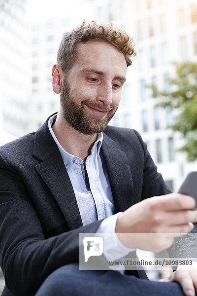 Lächelnder junger Geschäftsmann beim Blick auf das Handy im Freien
