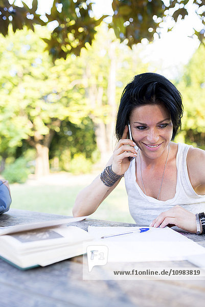 Lächelnde Frau sitzt im Park und telefoniert mit dem Smartphone.