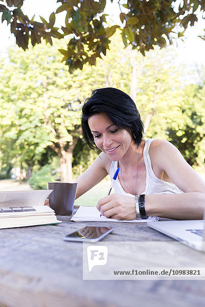 Lächelnde Frau sitzt am Tisch in einem Park und schreibt etwas auf.