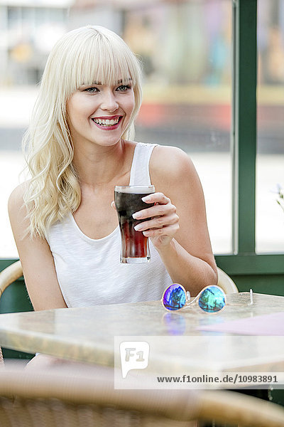 Porträt einer lächelnden blonden Frau  die auf dem Bürgersteig sitzt und Cola trinkt.