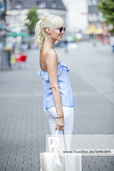 Lächelnde blonde Frau mit Tasche in einer Einkaufsstraße stehend