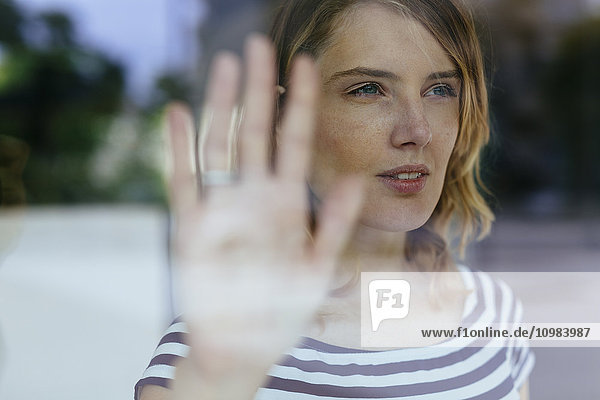 Porträt einer jungen Frau  die mit der Hand durchs Fenster blickt und Glas berührt